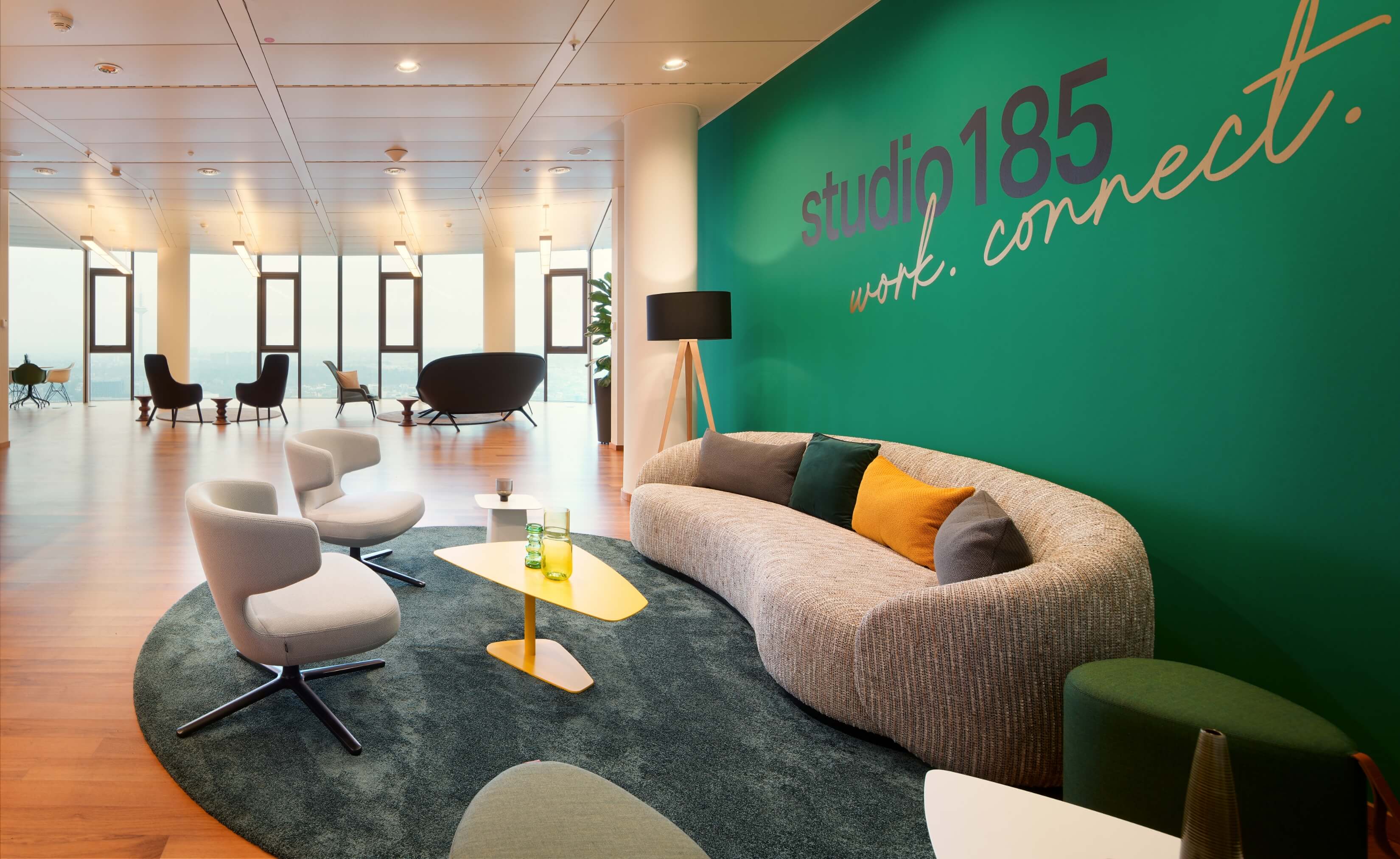 Eine Lounge mit Sofa und Sesseln auf Teppich vor einer grünen Wand, im Hintergrund weitere Sitzmöglichkeiten in einer großzügigen Raum in der 32. Etage des TOWER185